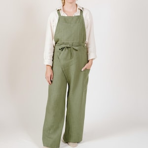 May include: Olive green linen jumpsuit with wide legs, a bib-style top, and a tie waist. The jumpsuit features two front pockets and adjustable shoulder straps. The model is wearing a white long-sleeve shirt underneath.