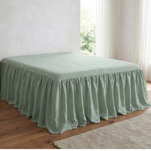 Puede incluir: Una falda de cama verde salvia con un diseño con volantes. La falda de cama cubre la base de la cama y tiene un estilo simple y elegante. La cama está en una habitación bien iluminada con una paleta de colores neutros.