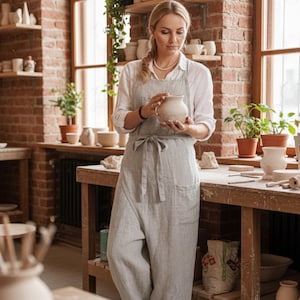 Puede incluir: Una mujer con un delantal gris claro sostiene un pequeño jarrón de cerámica sin terminar. El estudio presenta mesas de trabajo de madera, paredes de ladrillo y estantes que exhiben varias piezas de cerámica. Las plantas en macetas de terracota añaden un toque de vegetación.