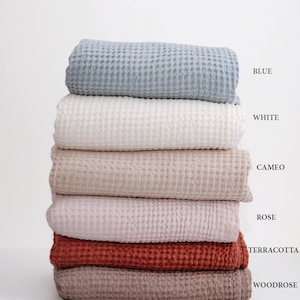 May include: A stack of six waffle weave towels in various colors, including blue, white, beige, pink, terracotta, and brown. The towels are folded and stacked on top of each other. The colors are labeled below the stack.