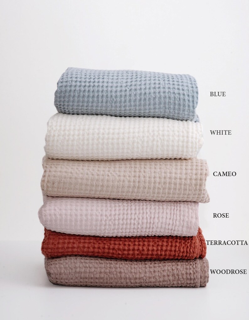 May include: A stack of six waffle weave towels in various colors, including blue, white, beige, pink, terracotta, and brown. The towels are folded and stacked on top of each other. The colors are labeled below the stack.