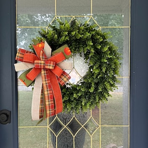 May include: A green boxwood wreath with a large orange, green, and brown plaid bow. The wreath is hanging on a dark blue door with a glass pane.