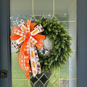 May include: A decorative wreath with a green, leafy base and a large bow. The bow features orange and white ribbons with football and Tennessee-themed designs. The wreath is hanging on a glass door with a dark blue frame.