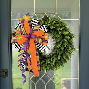 May include: A Halloween wreath with a green, leafy base and a large bow. The bow features orange, black, white, and purple ribbons with Halloween-themed patterns. The wreath is displayed on a glass door with a dark blue frame.