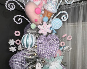 Puede incluir: Un árbol de Navidad decorado con un tema de dulces. La parte superior del árbol está adornada con un bastón de caramelo rosa y blanco, un cupcake y una piruleta azul y blanca. El árbol también está decorado con malla blanca y morada, copos de nieve blancos y un letrero que dice "Old Fashioned".