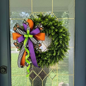 May include: A Halloween wreath with a green, leafy base and a large bow. The bow features orange, purple, black and white ribbons with Halloween-themed patterns and accents. The wreath is displayed on a glass door.