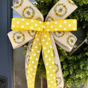May include: A yellow and beige bow with white polka dots and a bee and flower pattern. The bow is tied around a green wreath.