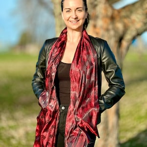 May include: A woman wearing a black leather jacket with a red and black patterned scarf. She is standing in front of a blurred background of trees.