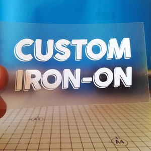 Può includere: Un foglio di trasferimento termoadesivo trasparente con la scritta "CUSTOM IRON-ON" in lettere bianche in grassetto. Il testo ha un effetto ombra e lo sfondo è un gradiente di blu. Una pallina da tennis è visibile sullo sfondo.