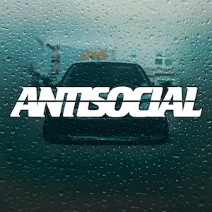Op de afbeelding: Een zwarte auto met een witte "ANTISOCIAL" sticker op de achterruit. De auto staat geparkeerd voor een gebouw met een wazige achtergrond van regendruppels op het raam.
