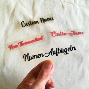 May include: Four iron-on labels with the text "Custom Name", "Mom Thermocollant", "Custom Name", and "Namen Aufb&uuml;geln".