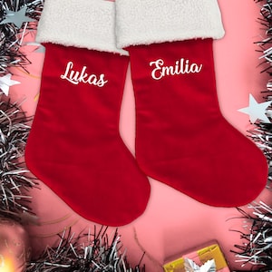 May include: Two red Christmas stockings with white faux fur trim. The stocking on the left is embroidered with the name "Lukas" and the stocking on the right is embroidered with the name "Emilia".