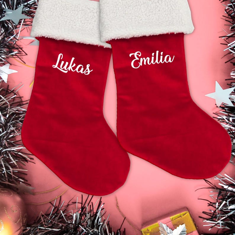 May include: Two red Christmas stockings with white faux fur trim. The stocking on the left is embroidered with the name "Lukas" and the stocking on the right is embroidered with the name "Emilia".