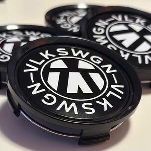 Peut inclure: Emblème Volkswagen noir et blanc avec le texte "VOLKSWAGEN" écrit en cercle autour de l'emblème. L'emblème est sur un enjoliveur en plastique noir.
