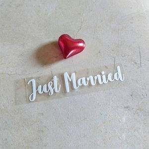 May include: A clear sticker with the text "Just Married" in white cursive lettering. A red heart-shaped decoration is above the sticker.