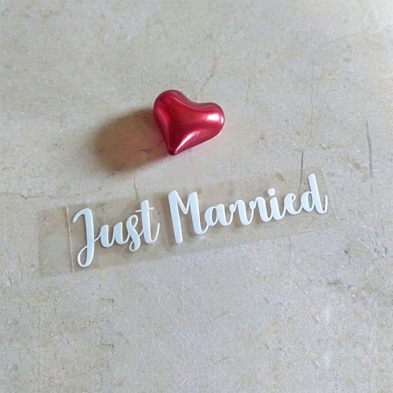 May include: A clear sticker with the text "Just Married" in white cursive lettering. A red heart-shaped decoration is above the sticker.