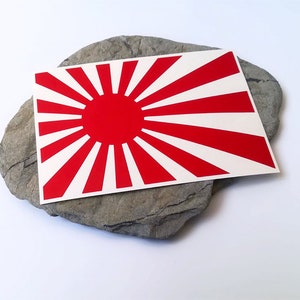 Puede incluir: Pegatina roja y blanca con el sol naciente sobre una roca gris. La pegatina es un diseño de bandera japonesa.