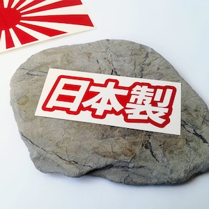 Puede incluir: Una pegatina rectangular con los caracteres japoneses "Hecho en Japón" en rojo, contorneados en blanco, se muestra sobre una piedra gris. Una bandera roja y blanca del sol naciente es visible en el fondo.