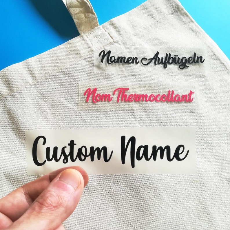 May include: A white tote bag with three iron-on labels. The labels are clear and have black text. The top label says "Namen Aufb&uuml;geln". The middle label says "Nom Thermocollant". The bottom label says "Custom Name".
