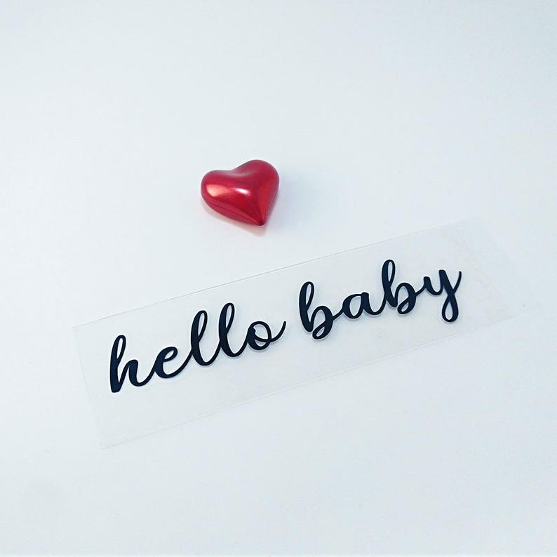 May include: A clear sticker with the text "hello baby" in black cursive lettering. A red heart is above the sticker.