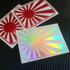 Könnte beinhalten: Zwei Vinyl-Aufkleber mit einem roten und weißen aufgehenden Sonnenmotiv. Ein Aufkleber ist ein Standard-Design in Rot und Weiß, während der andere ein holografischer Aufkleber mit einem Regenbogeneffekt ist.