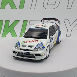 May include: A white and blue Ford Focus rally car model with Castrol and Michelin logos. The model has a rear spoiler and white wheels. The car is on a white platform with a green and white background.
