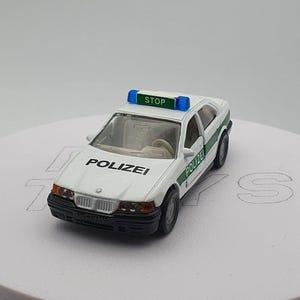 May include: A white toy police car with a green and blue light on top. The car has the word "POLIZEI" written on the side and "STOP" written on the light bar.
