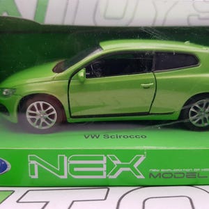 Puede incluir: Un coche modelo Volkswagen Scirocco verde en una caja verde y blanca. La caja tiene el texto "NEX MODELS" y "VW Scirocco" impreso en ella.