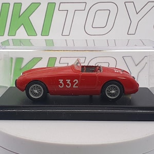 May include: A red toy car with the number 332 on the side, displayed in a clear plastic case. The car has silver wheels and a black base. The background has the text "RIKI TOYS".