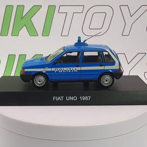 May include: A blue and white Fiat Uno police car model from 1987. The miniature car has the word "POLIZIA" printed on the side and sits on a black base. A small antenna and light are on the roof.