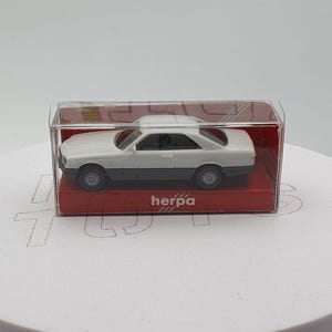 May include: A white and gray model car in a clear plastic display case. The car has a two-door coupe design. The display case sits on a red base with the brand name "herpa" printed on it.