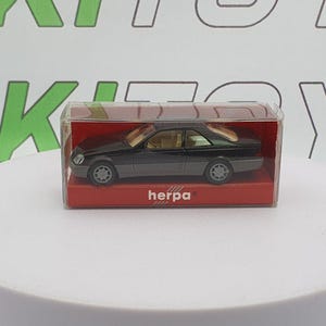 May include: A black and gray model car in a clear plastic case with a red base. The car has a coupe design with tan interior. The brand name "herpa" is printed on the base.