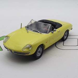 May include: A yellow toy car model of an Alfa Romeo convertible. The car has a black interior and a black soft top.
