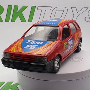 Op de afbeelding: Rood speelgoedautomodel van een Fiat Tipo 25 met gele en blauwe race strepen. De auto staat op een witte ronde displaystandaard.