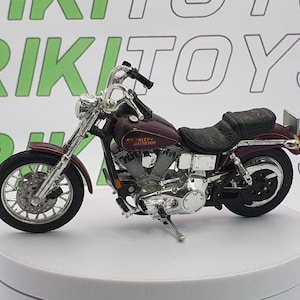 May include: A miniature burgundy Harley-Davidson motorcycle model with chrome accents. The model features a black seat, detailed engine components, and the Harley-Davidson logo. The motorcycle is displayed on a white surface.