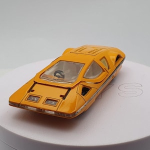 May include: A yellow toy car with a clear plastic windshield and a black steering wheel. The car has a futuristic design with a rounded body and a low profile.