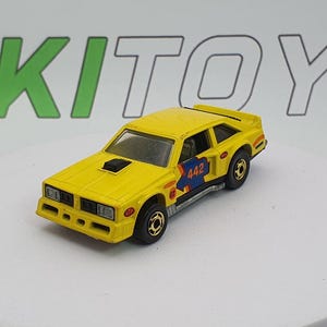 May include: A yellow die-cast toy car with black wheels. The car has a black hood scoop and blue and orange graphics with the number 442 on the side. The toy car is a vintage collectible.