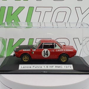 Könnte beinhalten: Ein rotes Modell eines Lancia Fulvia 1.6 HF RMC Rennwagens aus dem Jahr 1972 in einer transparenten Vitrine. Das Auto hat eine weiße Nummer "14" an der Seite und den Text "S. MUNARI-M. MANNUCCI" an der Seite. Das Auto steht auf einem schwarzen Sockel mit einem weißen Etikett, auf dem "Lancia Fulvia 1.6 HF RMC 1972" steht.
