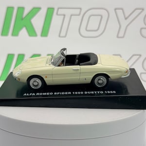 Può includere: Un'auto modello Alfa Romeo Spider 1600 Duetto 1966 color crema. La cabriolet in miniatura è su una base nera. L'auto ha interni neri e ruote argentate. Il testo "ALFA ROMEO SPIDER 1600 DUETTO 1966" è visibile.