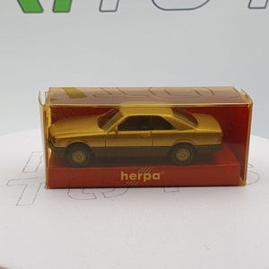 May include: A gold-colored model car in a clear plastic box with a red and white label that says "herpa".