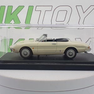 May include: A cream-colored model convertible car with a black interior, displayed in a clear plastic case. The miniature vehicle sits on a black base, showcasing its detailed design and classic aesthetic. The car's wheels are gray, and the model is a collectible item.