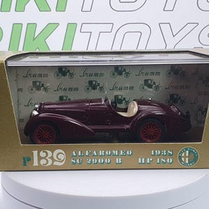 Könnte beinhalten: Ein weinrotes Modellauto Alfa Romeo SC 2900 B von 1938, in einer Box präsentiert. Das Modell hat rote Räder und eine beige Innenausstattung. Auf der Box steht "r139 ALFAROMEO SC 2900 B 1938 HP 180" und das Alfa Romeo Logo.