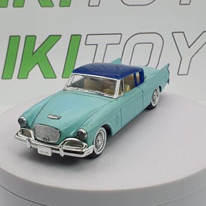 May include: A vintage teal and blue diecast model car. The car has a blue roof, white-walled tyres, and chrome detailing on the front bumper and grill. The model is displayed on a white rotating platform.