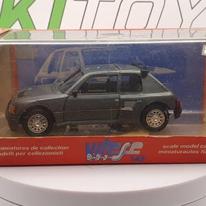 Peut inclure: Une maquette de voiture Peugeot 205 Turbo 16 gris foncé, présentée dans un boîtier en plastique transparent avec une boîte rouge et blanche. La boîte contient du texte en plusieurs langues, dont "Vitesse" et "scale model cars".