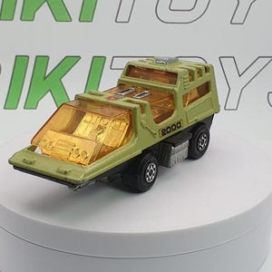 May include: A vintage olive green toy truck with transparent orange windows and the number "2000" printed on the side. The toy truck has black wheels and is set on a white surface.