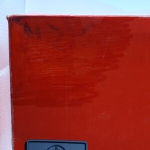 May include: A close-up of a red box with a black emblem at the bottom. The box has some scratches and markings on the surface. The box is against a white background.