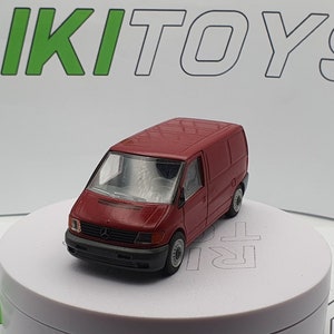 May include: A red toy van with a white front bumper and black headlights. The van has a white door and a black window. The van is sitting on a white surface.