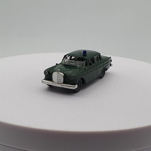 May include: A dark green die-cast model car with a silver front grill and a blue light on the roof. The classic car is a Mercedes-Benz, and it is displayed on a white surface.