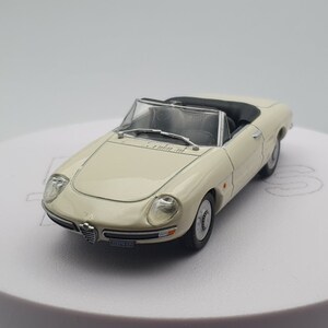 May include: A white diecast model car of an Alfa Romeo Spider, a classic Italian sports car. The car has a black convertible top and a black interior. The car is on a white surface.