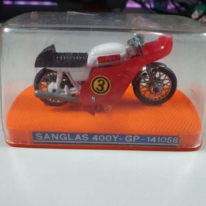 Puede incluir: Un modelo en miniatura de una motocicleta Sanglas 400Y roja, blanca y negra, encerrado en una caja de plástico transparente. El modelo tiene el número 3 y está sobre una base naranja. El texto 'SANGLAS 400Y-GP-141058' es visible.
